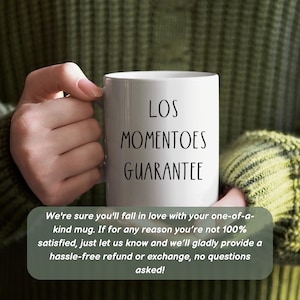May include: White ceramic mug with black text that reads "LOS MOMENTOES GUARANTEE". The mug is being held by a hand wearing a green sweater.