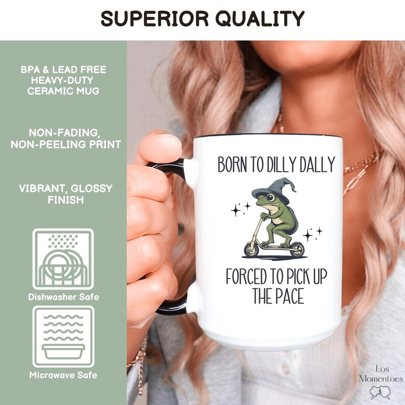 May include: White ceramic mug with a black handle. The mug has a green frog wearing a witch's hat riding a scooter. The text on the mug reads "Born to Dilly Dally" and "Forced to Pick Up the Pace".
