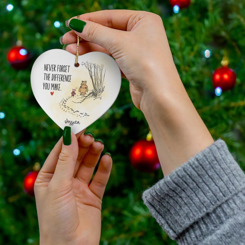 May include: A white heart-shaped ceramic ornament featuring a printed illustration of two characters walking in a forest with the text "NEVER FORGET THE DIFFERENCE YOU MAKE." and the name "Jessica" below, suspended by a gold-colored string.
