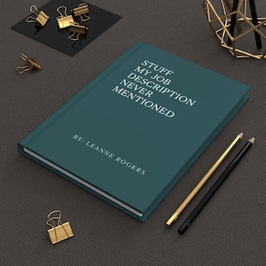 May include: A teal journal with the text "STUFF MY JOB DESCRIPTION NEVER MENTIONED" and "BY: LEANNE ROGERS" in white. Gold binder clips and pencils are scattered around the journal on a dark gray surface.
