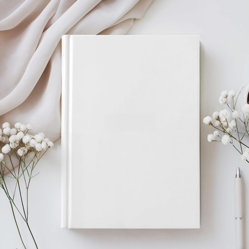 May include: A blank, white hardcover book with a clean, minimalist design. The book is positioned on a white surface, accompanied by delicate white flowers and a cream-colored pen. A soft, cream-colored fabric drapes in the background, creating a serene aesthetic.