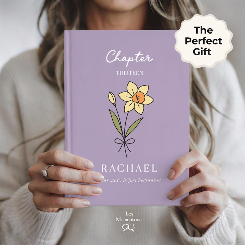 May include: A lavender-colored book with the words "Chapter Thirteen" and the name "Rachael" printed on the cover. A yellow daffodil illustration is also featured. The text "Our story is just beginning" is printed below the name.