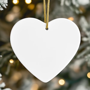 May include: A smooth, white, heart-shaped ornament with a small hole at the top for hanging. A thin, metallic gold ribbon is threaded through the hole. The text Los Momentoes is printed in white on the lower left side with a small icon below it.