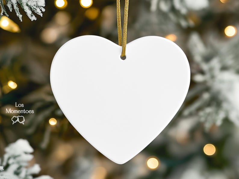 May include: A smooth, white, heart-shaped ornament with a small hole at the top for hanging. A thin, metallic gold ribbon is threaded through the hole. The text Los Momentoes is printed in white on the lower left side with a small icon below it.