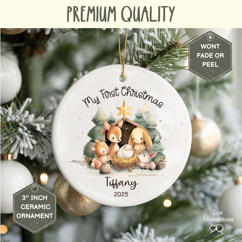 May include: A white ceramic Christmas ornament, 3 inches in diameter, with a watercolor illustration of a nativity scene. The ornament reads "My First Christmas", "Tiffany", and "2025". The ornament is hanging on a Christmas tree.