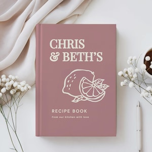 May include: A dusty rose hardcover recipe book featuring the white text CHRIS & BETH'S, a line art illustration of a lemon and a lemon slice, and the text RECIPE BOOK from our kitchen with love printed on the front cover.
