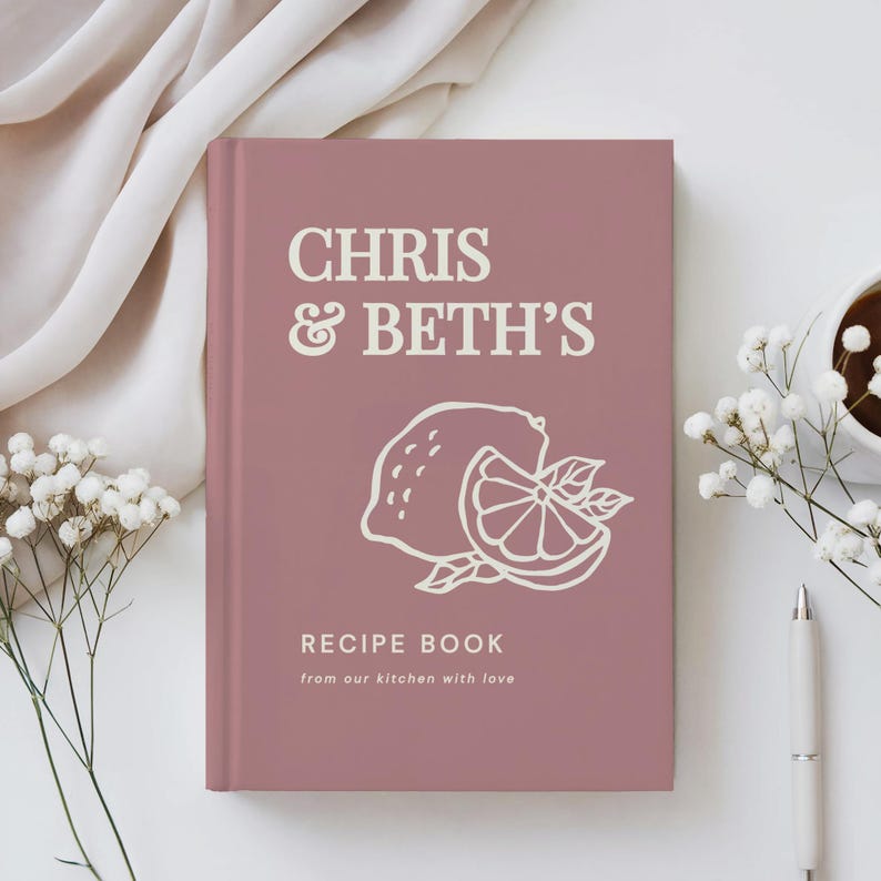 May include: A dusty rose hardcover recipe book featuring the white text CHRIS & BETH'S, a line art illustration of a lemon and a lemon slice, and the text RECIPE BOOK from our kitchen with love printed on the front cover.