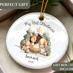 May include: White ceramic Christmas ornament with a nativity scene illustration. The ornament features the text "My First Christmas," a name, and the year 2025. Includes a gold string for hanging. The ornament is in a gift box.