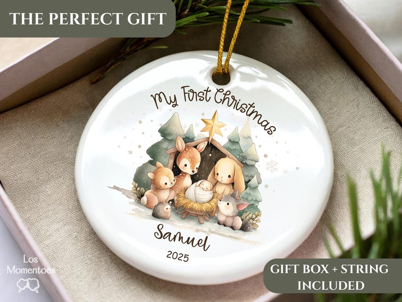 May include: White ceramic Christmas ornament with a nativity scene illustration. The ornament features the text "My First Christmas," a name, and the year 2025. Includes a gold string for hanging. The ornament is in a gift box.