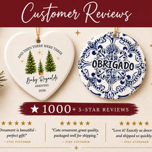 May include: Two ceramic ornaments, one heart-shaped and one circular. The heart ornament features three green Christmas trees, gold stars, and text reading AND THEN THERE WERE THREE Baby Reynolds ARRIVING 2026. The circular ornament has a blue and white floral pattern and the word OBRIGADO.