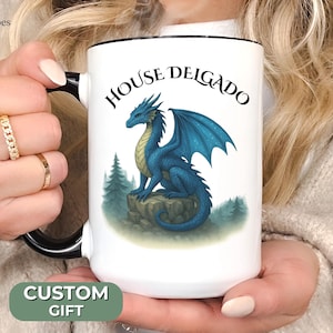 May include: White ceramic mug with a black rim and handle. The mug features a blue dragon with wings and the text "House Delgado" in black.