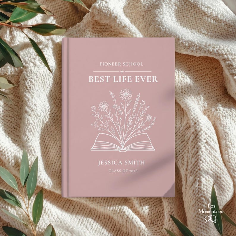 May include: A dusty rose-colored journal with the text "Pioneer School BEST LIFE EVER" and the name "Jessica Smith, Class of 2026." A white floral illustration is above an open book design. The journal rests on a cream-colored knitted blanket.