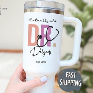 May include: White insulated tumbler with a handle, featuring the text "Actually it's DR. Delgado" and a stethoscope graphic. The tumbler has a clear lid and a silver rim. The text "EST 2025" is also printed on the tumbler.