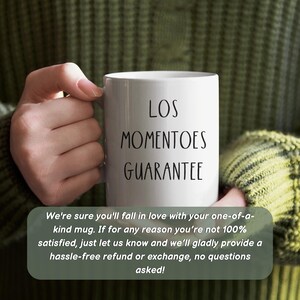 May include: A white ceramic mug held by a person, with the words "LOS MOMENTOES GUARANTEE" in black. The mug is held against a green sweater. The text below the mug reads: "We're sure you'll fall in love with your one-of-a-kind mug."