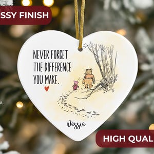 May include: A white heart-shaped ceramic ornament with a glossy finish, featuring a gold hanging cord. The design includes a sketch of Winnie the Pooh and Piglet, the text "NEVER FORGET THE DIFFERENCE YOU MAKE.", a small red heart, and the name "Jessie".