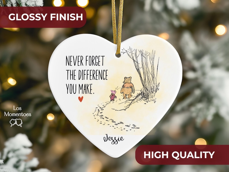May include: A white heart-shaped ceramic ornament with a glossy finish, featuring a gold hanging cord. The design includes a sketch of Winnie the Pooh and Piglet, the text "NEVER FORGET THE DIFFERENCE YOU MAKE.", a small red heart, and the name "Jessie".