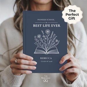 May include: A navy blue journal with the text "PIONEER SCHOOL BEST LIFE EVER" and the name "REBECCA" with "CLASS OF 2026". The cover features a floral design above an open book illustration. A white sticker says "The Perfect Gift".