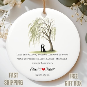 May include: A white ceramic ornament with a willow tree illustration and a couple standing beneath it. Text includes a quote about strength and the names "Elysia" and "Tyler" with a date. A jute string is attached for hanging.