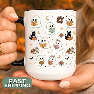 May include: White ceramic mug with a black handle featuring a repeating pattern of cartoon ghosts reading books, black cats, and bats. The mug has a green "FAST SHIPPING" sticker on the bottom. The mug is held by a person.