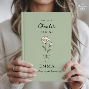 May include: A sage green journal with the words "THE NEXT CHAPTER BEGINS" and the name "EMMA" printed on the cover. A daisy illustration is featured below the text. The phrase "story is just beginning" is printed at the bottom.