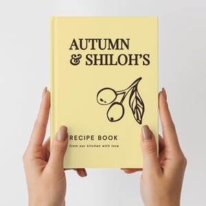 May include: A pale yellow hardcover recipe book featuring dark brown text that reads AUTUMN & SHILOH'S RECIPE BOOK from our kitchen with love. A simple dark brown line drawing of two olives on a branch with a single leaf is centered on the cover.