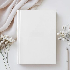 May include: A blank white hardcover book positioned vertically. The book is surrounded by delicate white baby's breath flowers and a sleek white pen. A soft, draped beige fabric is visible in the upper left corner of the composition.