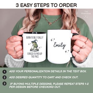 May include: Two white ceramic mugs with black rims. One mug has a green frog wearing a witch's hat riding a scooter with the text "Born to Dilly Dally, Forced to Pick Up the Pace". The other mug has the name "Emily" written on it.