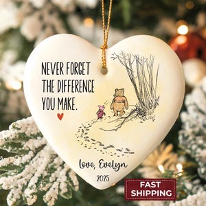 May include: A heart-shaped white ceramic ornament featuring a black ink illustration of Winnie the Pooh and Piglet walking away. The text reads: NEVER FORGET THE DIFFERENCE YOU MAKE. Love, Evelyn 2025. A small red heart and gold hanging string.