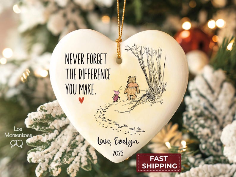 May include: A heart-shaped white ceramic ornament featuring a black ink illustration of Winnie the Pooh and Piglet walking away. The text reads: NEVER FORGET THE DIFFERENCE YOU MAKE. Love, Evelyn 2025. A small red heart and gold hanging string.