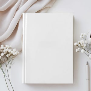 May include: A plain white hardback book dominates the image, accompanied by a white pen, sprigs of white flowers, and a beige fabric. The composition is clean and minimalist, suggesting a focus on writing or journaling.
