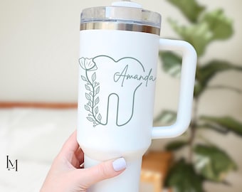 May include: A white insulated tumbler with a handle and a clear lid, featuring a sage green line art graphic of a tooth decorated with a floral vine on the left side and the name Amanda written in a cursive script on the right side of the tooth.