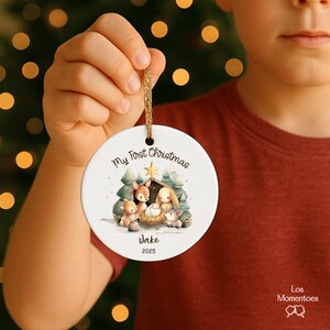 May include: A white, round Christmas ornament with a gold string. The ornament features a watercolor illustration of a nativity scene with animals and the text "My First Christmas", the name "Jake", and the year "2025".