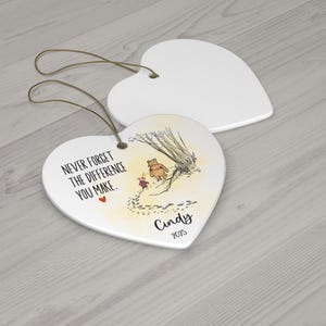 May include: Two heart-shaped white ceramic ornaments with gold hanging strings. One ornament features a printed illustration of Winnie the Pooh and Piglet, the text "NEVER FORGET THE DIFFERENCE YOU MAKE.", a small red heart, the name "Cindy", and "2025".