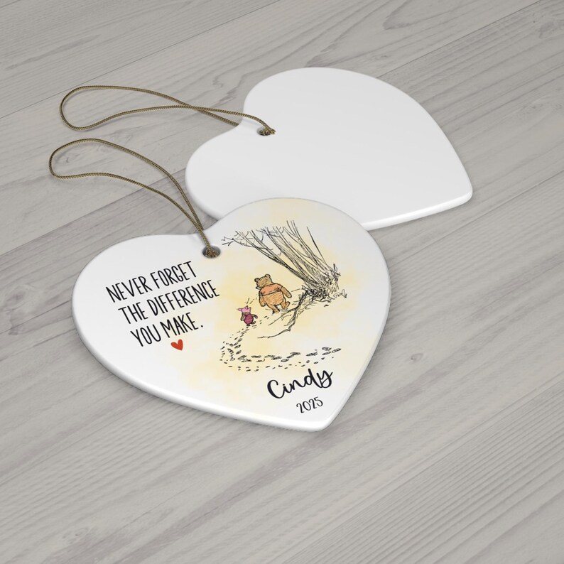 May include: Two heart-shaped white ceramic ornaments with gold hanging strings. One ornament features a printed illustration of Winnie the Pooh and Piglet, the text "NEVER FORGET THE DIFFERENCE YOU MAKE.", a small red heart, the name "Cindy", and "2025".