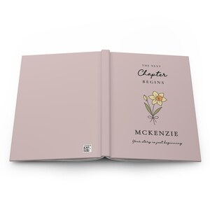 May include: Open journal with a pale pink cover. The right page features the text "THE NEXT Chapter BEGINS" above a daffodil illustration and the name "MCKENZIE". The phrase "Your story is just beginning" is below.