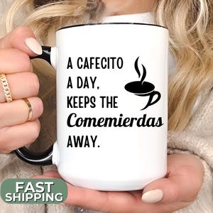 May include: White ceramic mug with a black rim and handle. The mug has black text that reads "A cafecito a day, keeps the comemierdas away." There is a black illustration of a steaming cup of coffee.