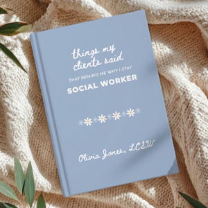 May include: A light blue journal with the text "things my clients said" and "SOCIAL WORKER" in white. The journal has a daisy chain design and the name "Olivia Jones, LCSW" at the bottom. The journal is lying on a cream-colored knitted blanket.