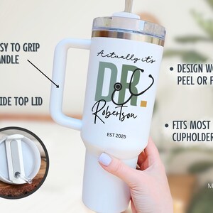 May include: White insulated tumbler with a handle, featuring the text "Actually it's DR Robertson EST 2025" and a stethoscope graphic. The tumbler has a slide top lid and is designed to fit most cupholders.