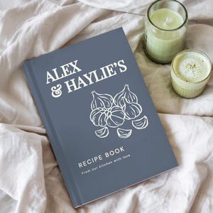 May include: A slate blue hardcover recipe book with white text that reads ALEX & HAYLIE'S RECIPE BOOK From our kitchen with love. The cover features a white line drawing of garlic bulbs and cloves centered on the front.