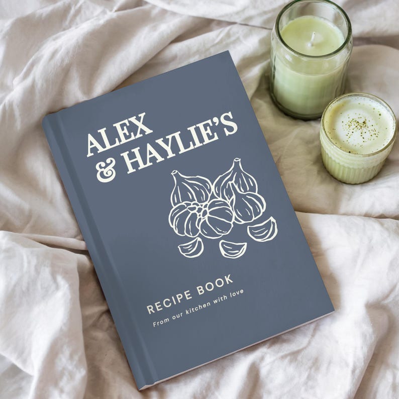 May include: A slate blue hardcover recipe book with white text that reads ALEX & HAYLIE'S RECIPE BOOK From our kitchen with love. The cover features a white line drawing of garlic bulbs and cloves centered on the front.