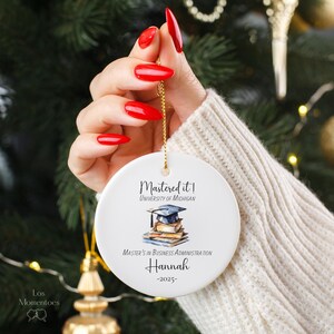 May include: White ceramic ornament with a watercolor illustration of a graduation cap on top of a stack of books. The text reads "Mastered it! University of Michigan Master's in Business Administration Hannah -2025- "