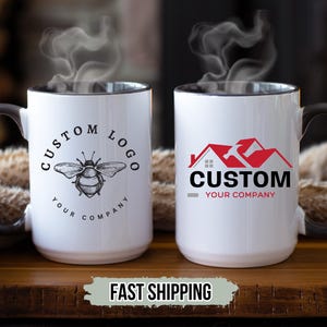 May include: Two white ceramic mugs with black handles and rims. One mug features a bee graphic with the text "CUSTOM LOGO YOUR COMPANY". The other mug has a red house and hammer graphic with the text "CUSTOM YOUR COMPANY". Steam rises from the mugs.