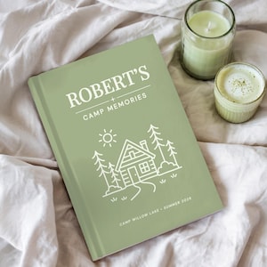 May include: A sage green hardcover journal titled "ROBERT'S CAMP MEMORIES" with a cabin illustration. The journal includes the text "CAMP WILLOW LAKE &bull; SUMMER 2026". A lit candle and a small cup of matcha are next to the journal.
