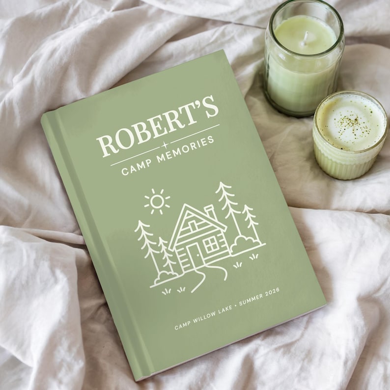 May include: A sage green hardcover journal titled "ROBERT'S CAMP MEMORIES" with a cabin illustration. The journal includes the text "CAMP WILLOW LAKE &bull; SUMMER 2026". A lit candle and a small cup of matcha are next to the journal.
