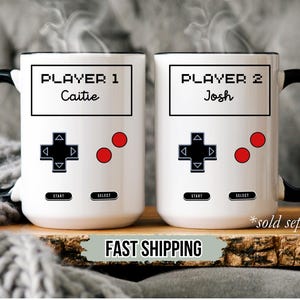 K&ouml;nnte beinhalten: Zwei wei&szlig;e Keramikbecher mit schwarzen Griffen und R&auml;ndern, die im Design eines Videospiel-Controllers gestaltet sind. Auf einem Becher steht "PLAYER 1 Caitie", auf dem anderen "PLAYER 2 Josh". Jeder Becher hat ein Steuerkreuz, zwei rote Kn&ouml;pfe und "START"- und "SELECT"-Tasten.