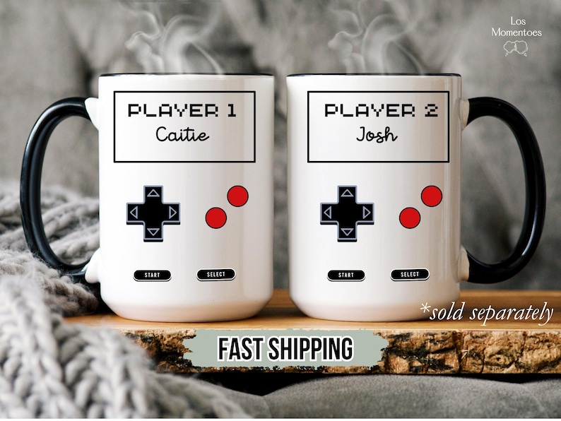 K&ouml;nnte beinhalten: Zwei wei&szlig;e Keramikbecher mit schwarzen Griffen und R&auml;ndern, die im Design eines Videospiel-Controllers gestaltet sind. Auf einem Becher steht "PLAYER 1 Caitie", auf dem anderen "PLAYER 2 Josh". Jeder Becher hat ein Steuerkreuz, zwei rote Kn&ouml;pfe und "START"- und "SELECT"-Tasten.