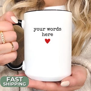 May include: White ceramic mug with a black handle and rim. The mug features the text "your words here" above a small red heart. The mug is held by a person.