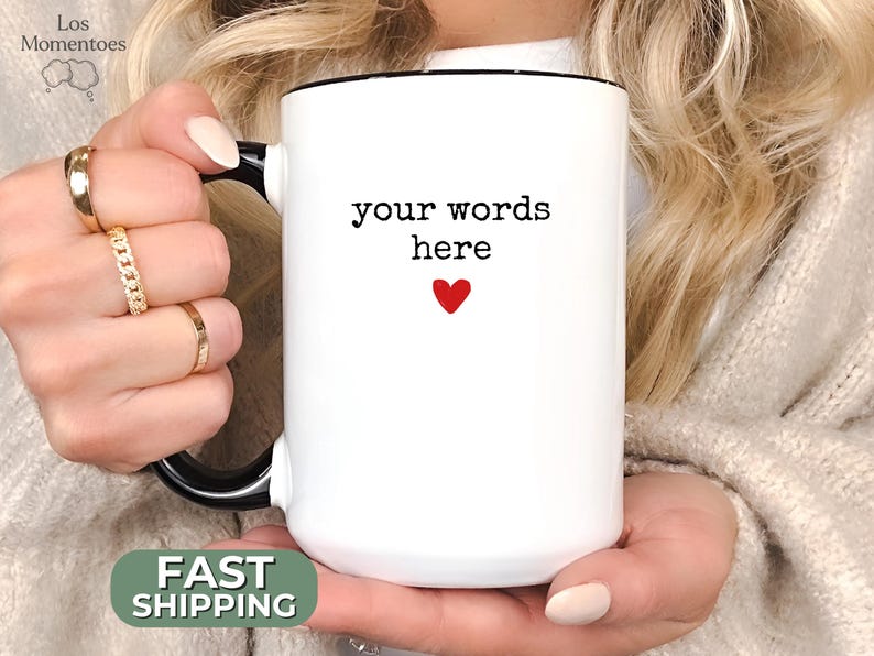 May include: White ceramic mug with a black handle and rim. The mug features the text "your words here" above a small red heart. The mug is held by a person.