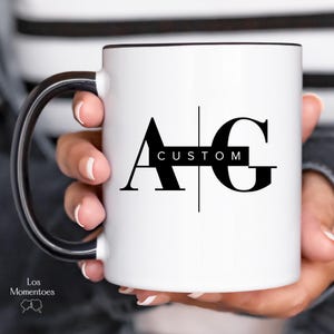 May include: White ceramic mug with a black rim and handle. The mug features the letters "A" and "G" in a bold, black font, with the word "CUSTOM" in a black rectangle between them. The mug is held by a person.