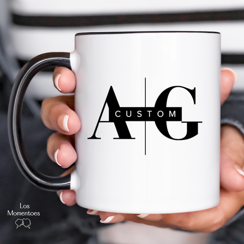 May include: White ceramic mug with a black rim and handle. The mug features the letters "A" and "G" in a bold, black font, with the word "CUSTOM" in a black rectangle between them. The mug is held by a person.
